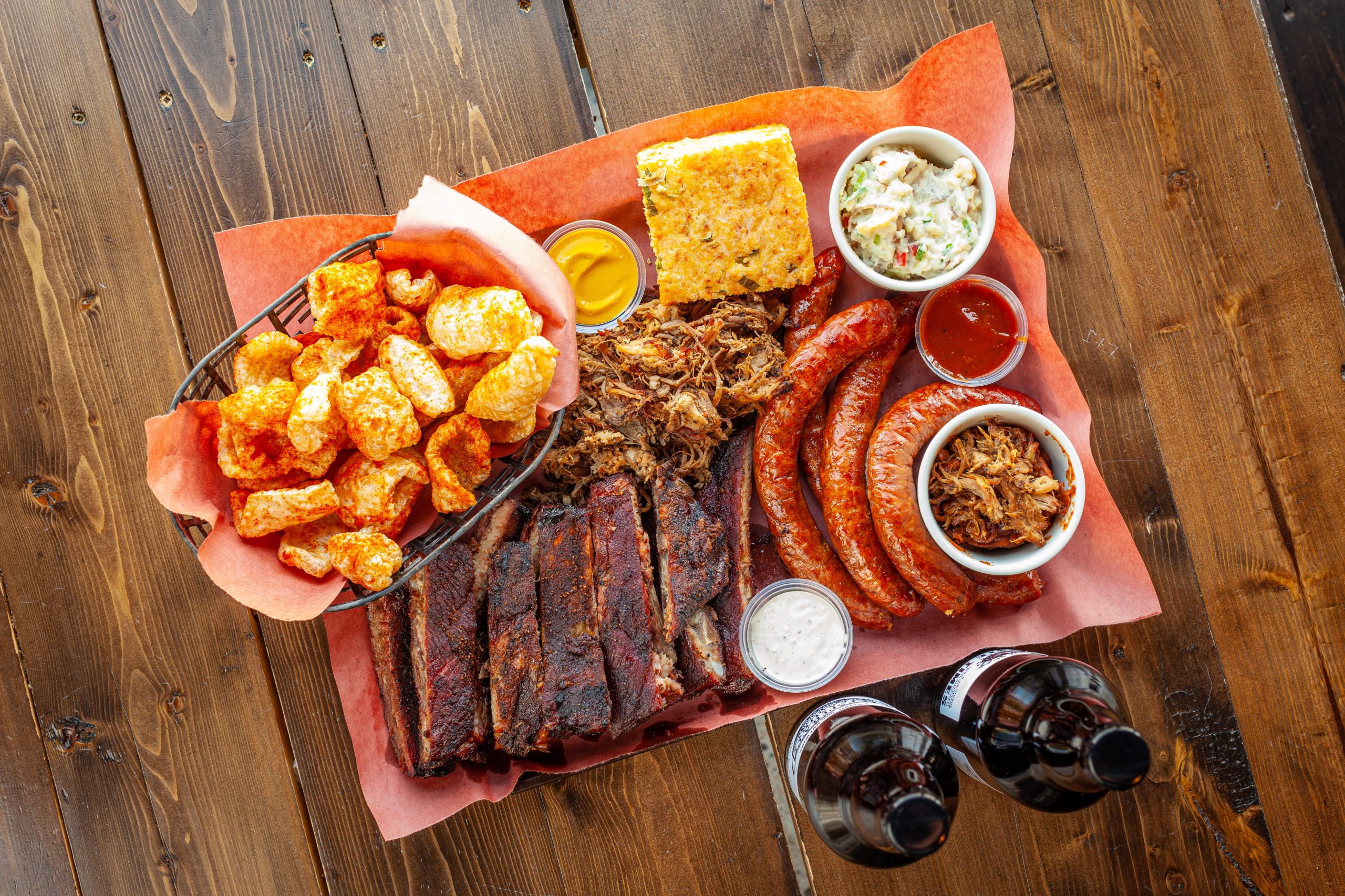 A delicious arrangement of barbecued meats, sides and barbecue sauces on a large tray with butcher paper on a wood table, with two Prairie Dog special release bottled beers.