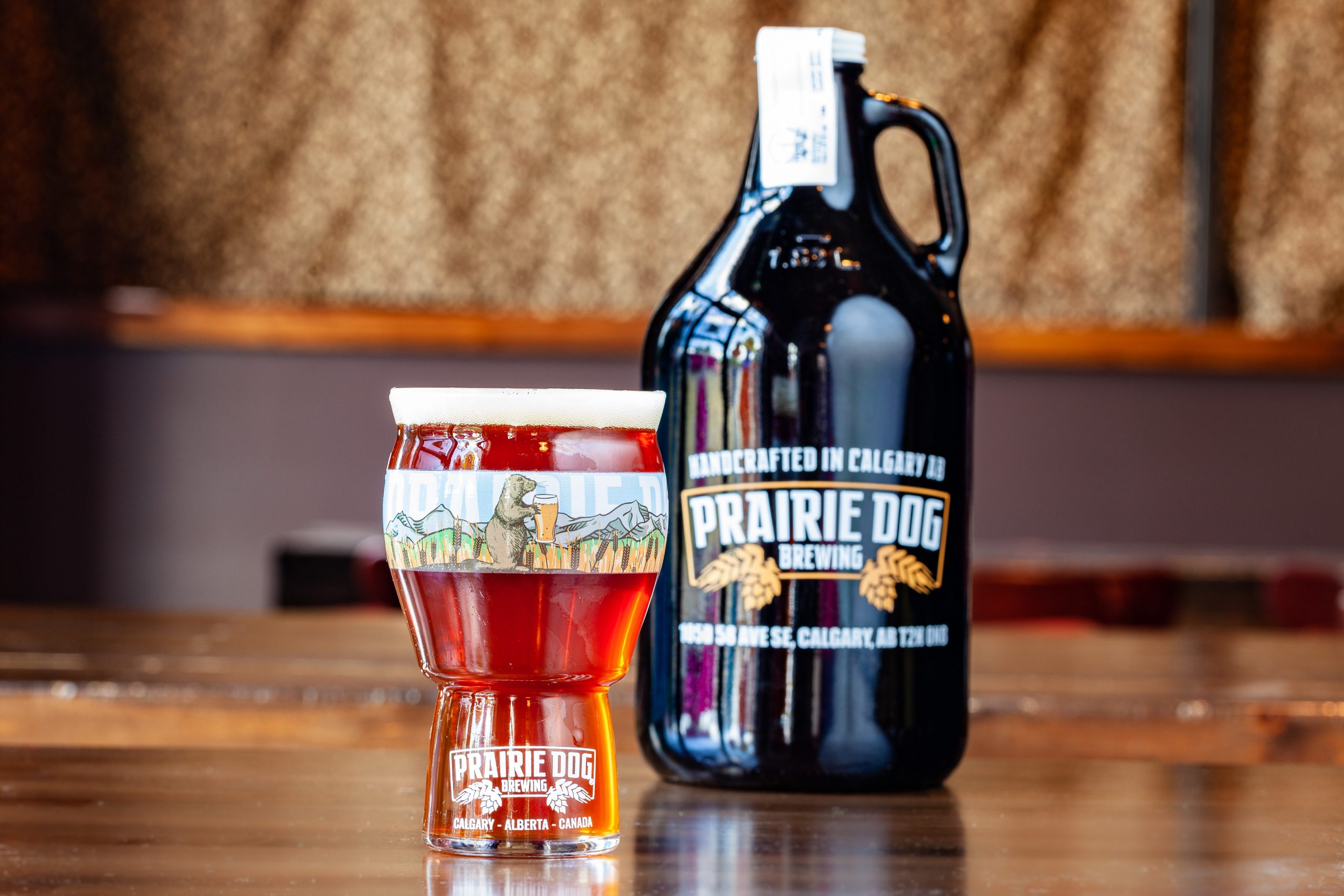 Prairie Dog Brewing's Alby the Elder Double IPA (DIPA) in a branded 16-oz US pint glass with a 64-oz branded growler in the background.