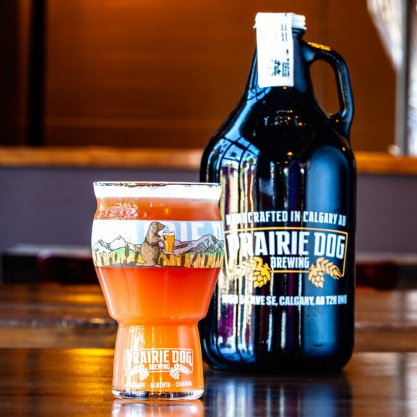 Prairie Dog Brewing and Hubtown Brewery's Berry Manly collaboration fruited Belgian-style blonde ale in a branded 16-oz US pint glass with a 64-oz branded growler in the background.