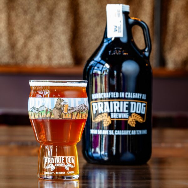 Prairie Dog Brewing's Jitter Bear Coffee Kolsch with Rebel Bean coffee, in a branded 16-oz US pint glass with a 64-oz growler jug in the background.