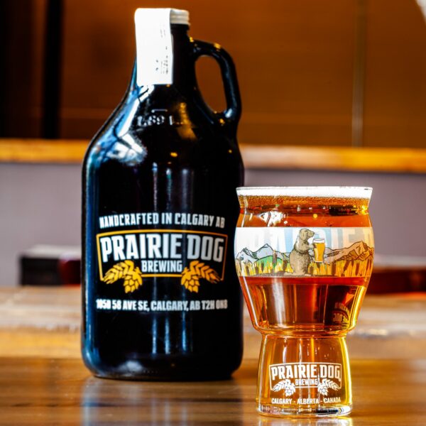 Prairie Dog Brewing's Super B Wheat (sorry, the batch pictured was not hazy), in a 16-oz US pint glass with a branded 64-oz growler jug in the background.