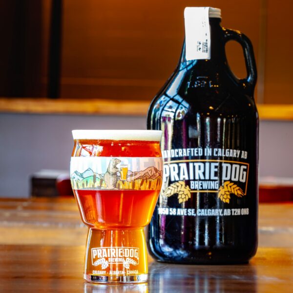 Prairie Dog Brewing's Tail Twitcher IPA craft beer in a branded 16-oz US pint glass, with a 64-oz branded growler jug in the background.