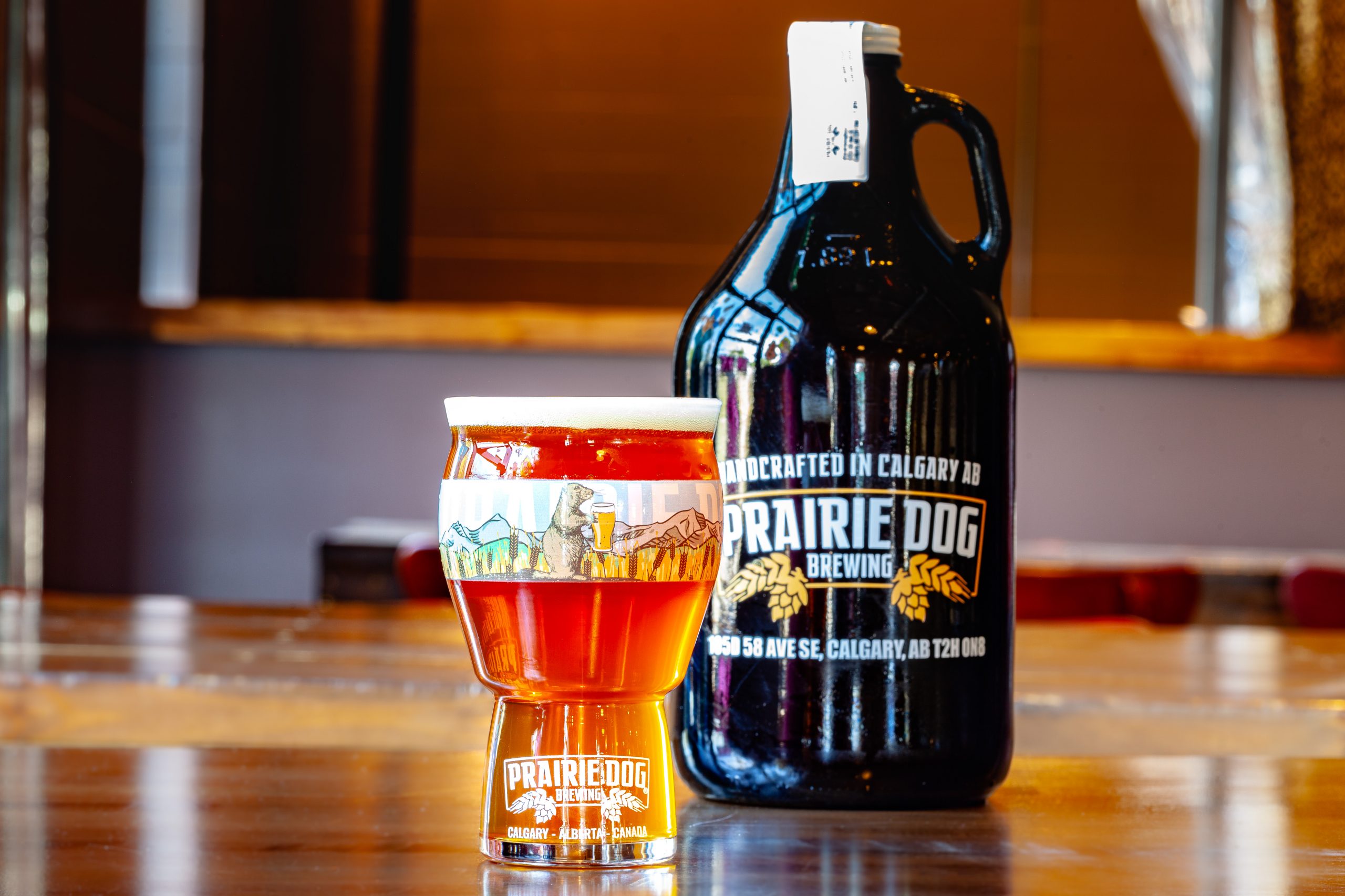 Prairie Dog Brewing's Tail Twitcher IPA craft beer in a branded 16-oz US pint glass, with a 64-oz branded growler jug in the background.