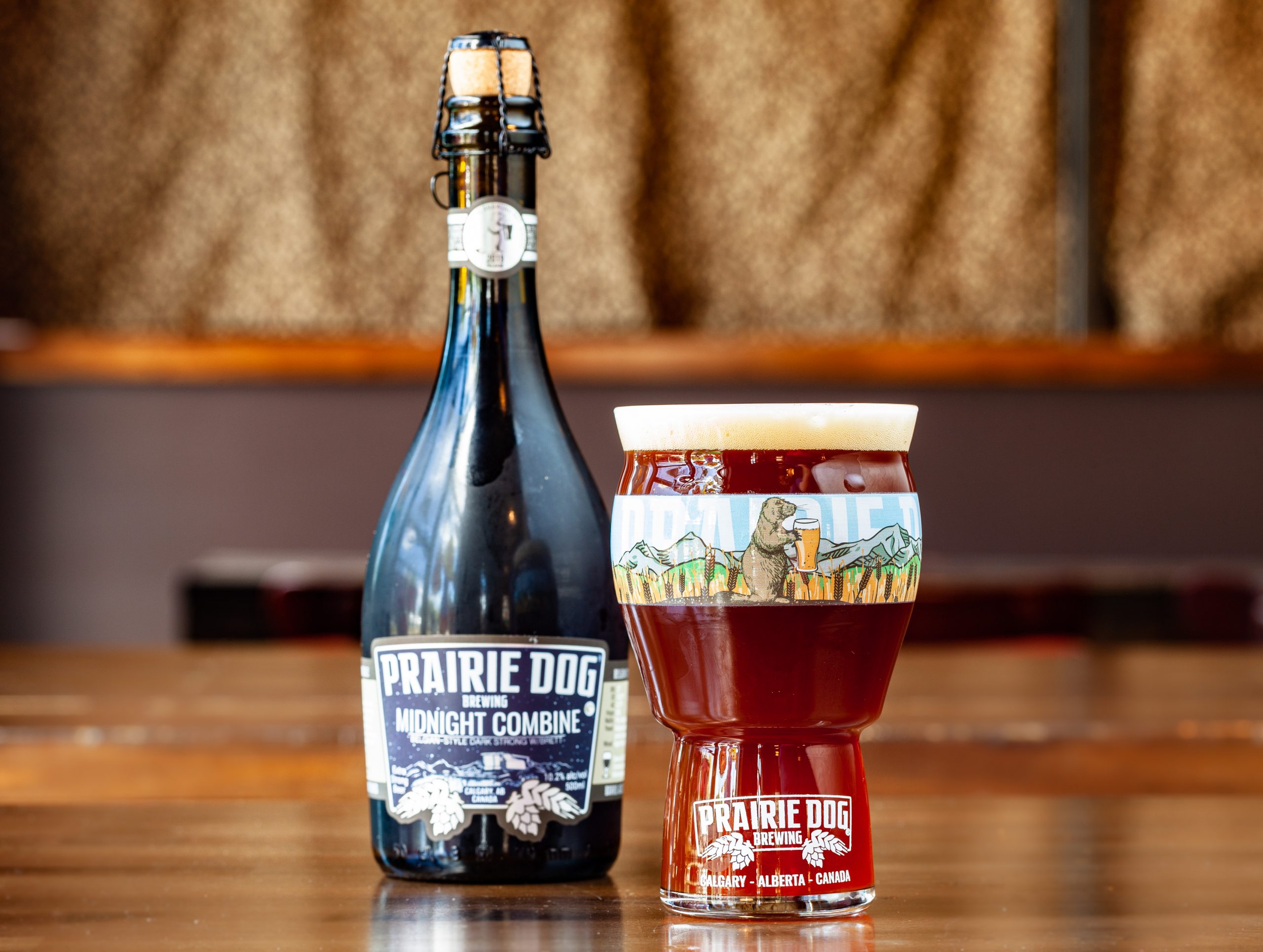 A 16-oz glass of Prairie Dog Brewing Midnight Combine barrel-aged Belgian-style Dark Strong Ale in a 16-oz branded pint glass, with a 500-mL bottle.