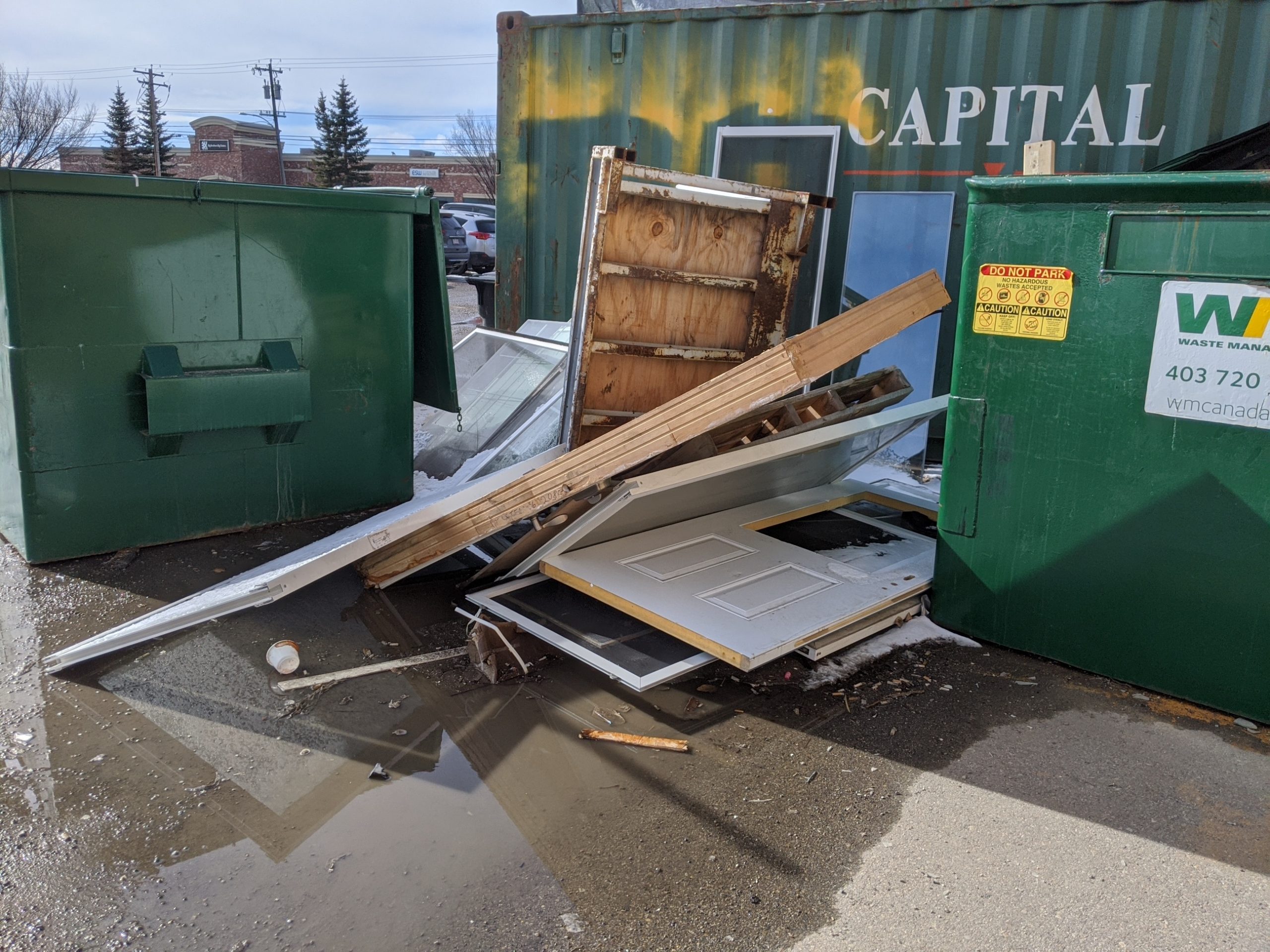 Image of a large pile of broken doors and old window glass piled up between garbage bins and Prairie Dog Brewing's green sea can in their back parking area.