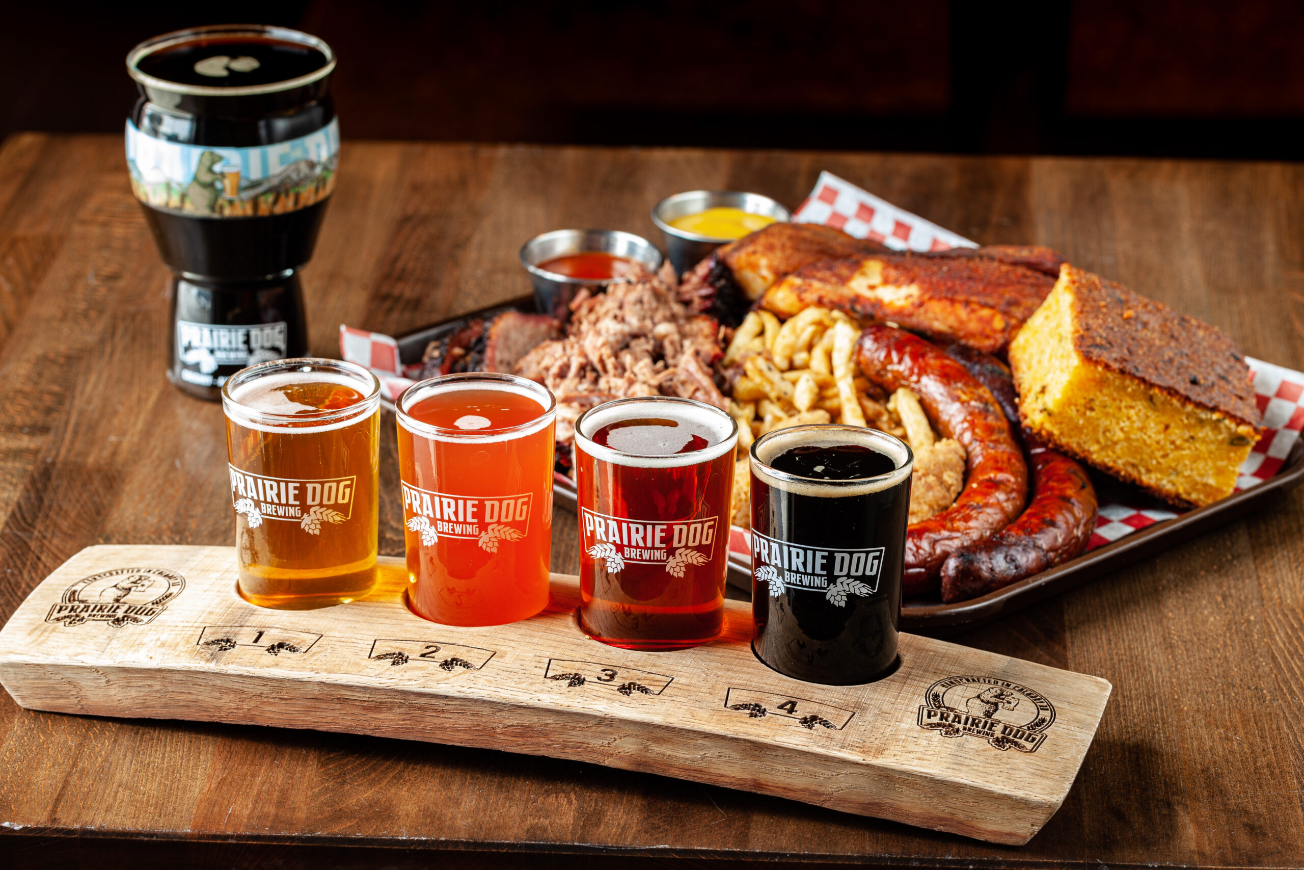 A Prairie Dog Brewing beer flight in the foreground with a Coterie barbecue platter and pint of stout in the rear, on a tabletop.