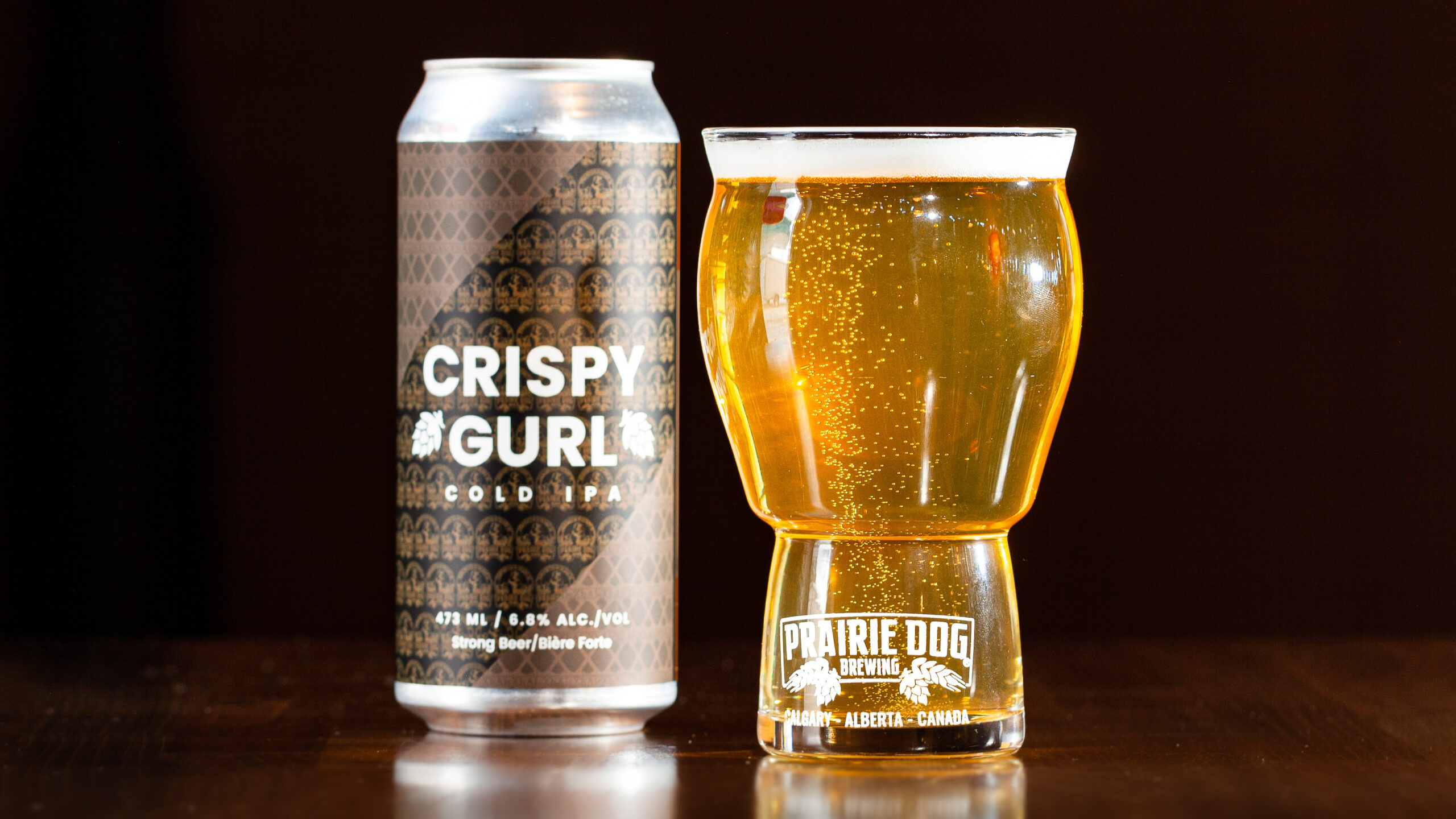 Prairie Dog Brewing and XhAle Brew Co. collab beer Crispy Gurl, a Cold IPA, in a 473mL can beside a pour of the same beer in a Prairie Dog Brewing beer glass.