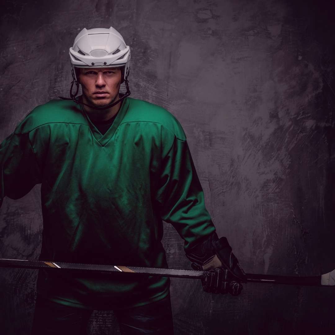 A hockey player in a green jersey stands holding his stick on a grey backdrop.