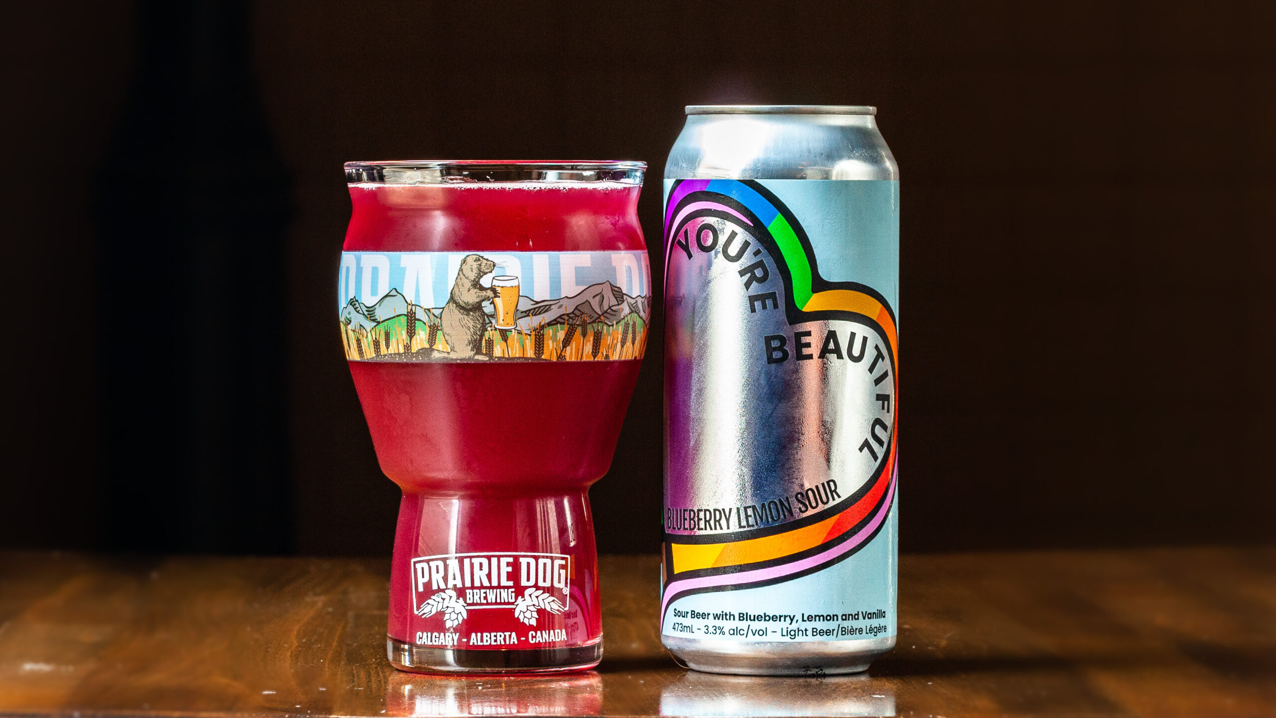 A 473-ml pint glass of brightly coloured beer next to a can of Prairie Dog Brewing You're Beautiful Blueberry Lemon Sour Beer in full colour wrapped label.