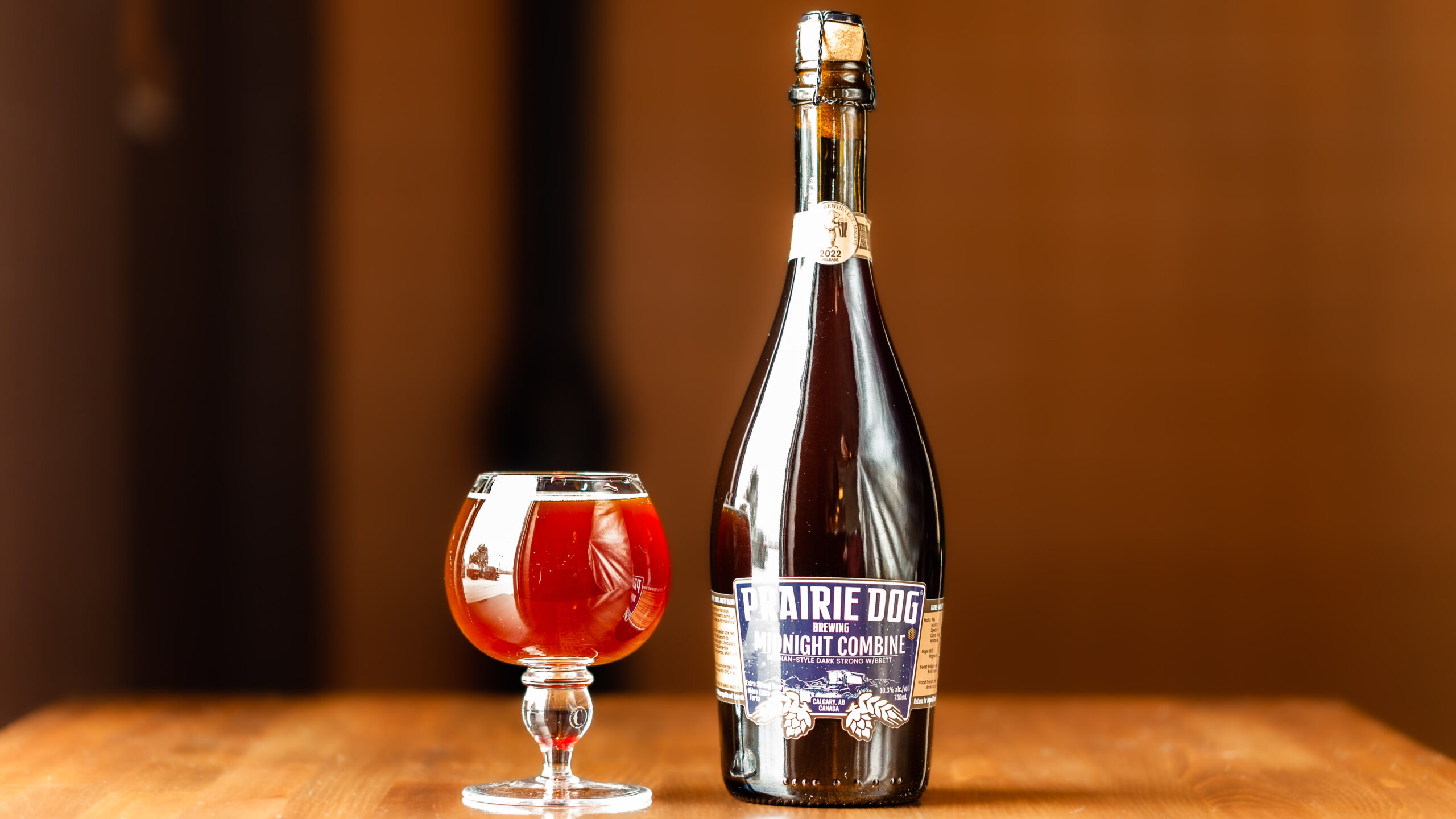 Prairie Dog Brewing's 2022 Midnight Combine Bottle and Tulip A dark, high-contrast image of oak whiskey and wine barrels stacked 3-high on racks at Prairie Dog Brewing.