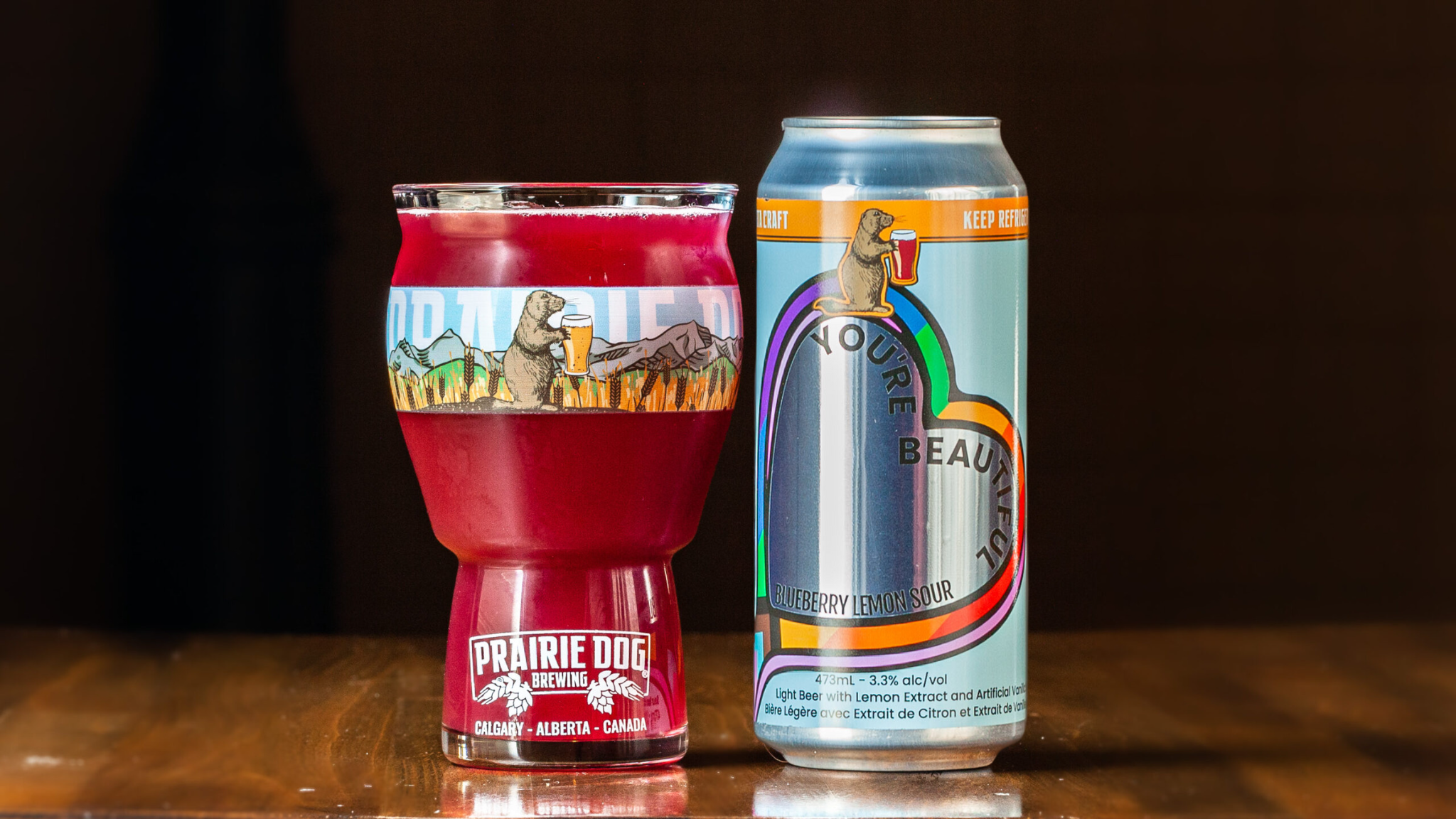 A 473-ml pint glass of brightly coloured beer next to a can of Prairie Dog Brewing You're Beautiful Blueberry Lemon Sour Beer in full colour wrapped label.