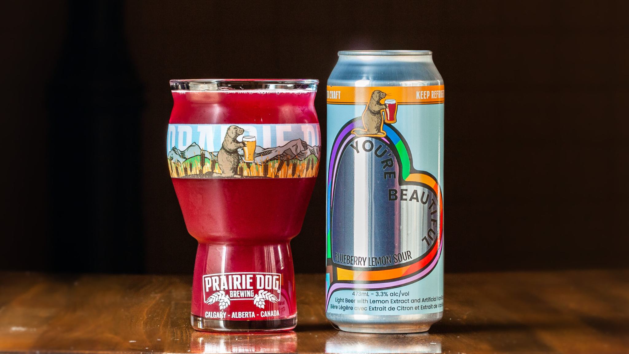 You're Beautiful Can + Glass A 473-ml pint glass of brightly coloured beer next to a can of Prairie Dog Brewing You're Beautiful Blueberry Lemon Sour Beer in full colour wrapped label.